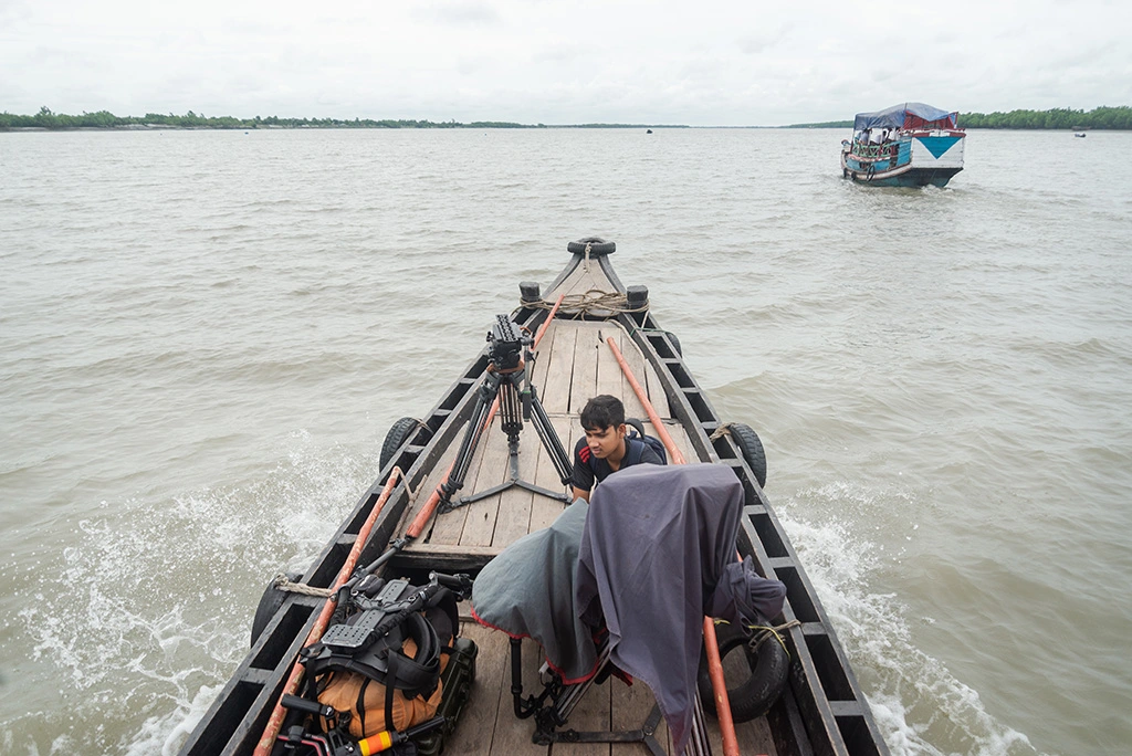 A Bangladeshi film fixer negotiating transport logistics at a busy ferry terminal, holding a phone and paperwork.